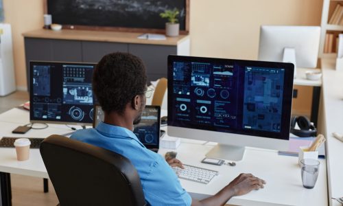 Rear view of young African American businessman sitting by workplace in front of desktop computers with graphic data on screens in office